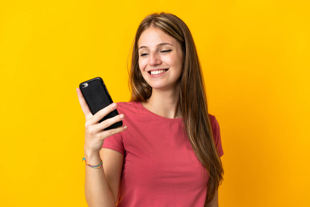 Mujer joven usando teléfono móvil aislado sobre fondo amarillo con expresión feliz - Foto, Imagen