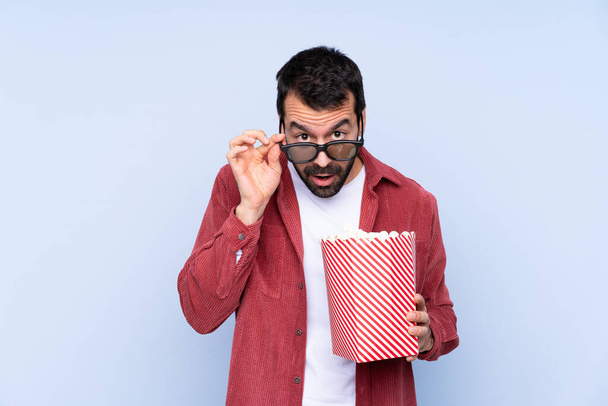 Young caucasian man over blue background surprised with 3d glasses and holding a big bucket of popcorns - Photo, Image