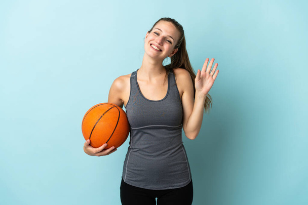 Junge Frau beim Basketballspielen auf blauem Hintergrund salutiert mit glücklicher Miene mit der Hand - Foto, Bild