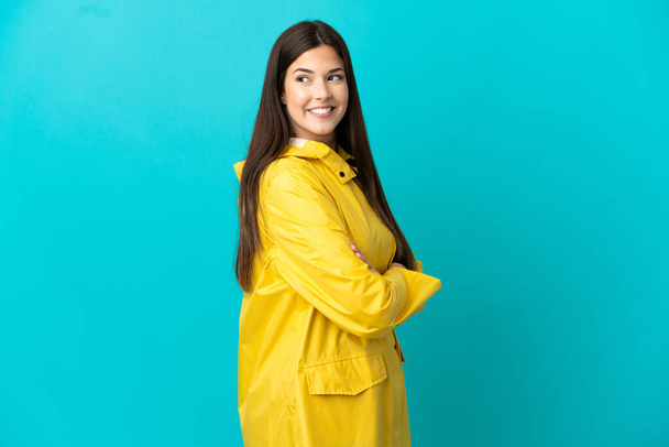 Teenager Brazilian girl wearing a rainproof coat over isolated blue background with arms crossed and happy - Photo, Image