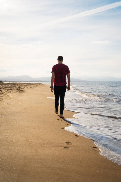 A Caucasian man is walking along the seashore. It is a sunny day and the sea is calm. He is wearing a red short-sleeved T-shirt and black jeans. He is alone on the beach. Les Marines, Denia. - Foto, immagini