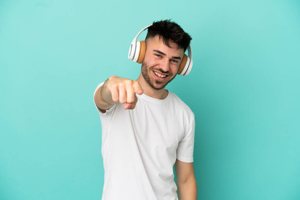 Young caucasian man isolated on blue background listening music and pointing to the front - Photo, Image