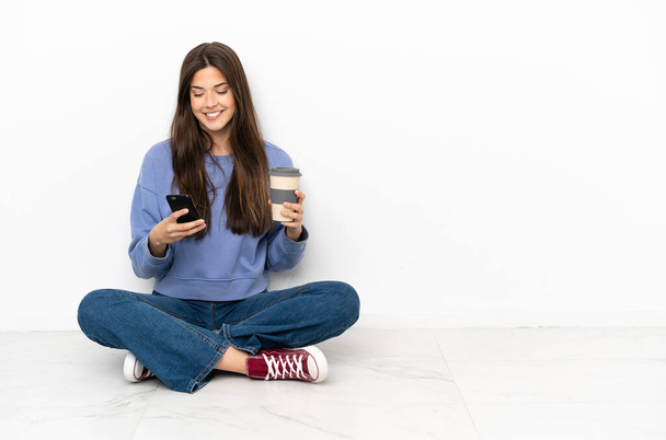 Jeune femme assise sur le sol tenant du café à emporter et un mobile - Photo, image