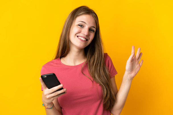 黄色の背景に孤立した携帯電話を使用して若い女性幸せな表情で手で塩漬け - 写真・画像