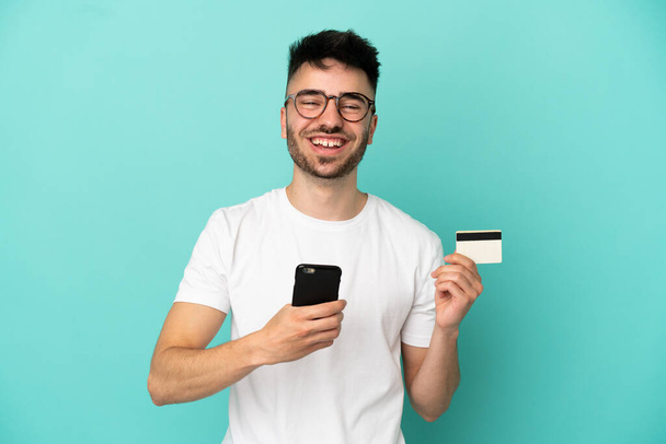 Young caucasian man isolated on blue background buying with the mobile with a credit card - 写真・画像