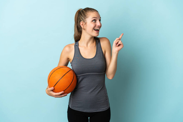 Junge Frau spielt Basketball isoliert auf blauem Hintergrund in der Absicht, die Lösung zu realisieren, während sie einen Finger hebt - Foto, Bild