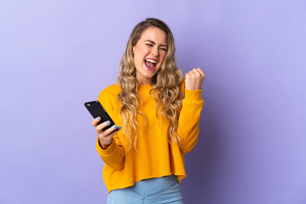 Young Brazilian woman isolated on purple background with phone in victory position - Fotó, kép