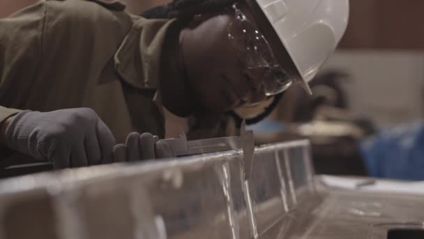 Medium close-up of female African worker wearing white hard hat and safety goggles, leaning forward, using caliper measuring width of metallic piece - Footage, Video