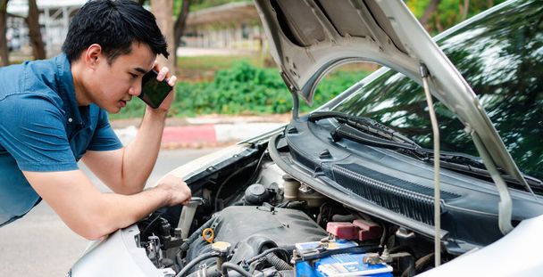 Angry Asian man and using mobile phone calling for assistance after a car breakdown on street. Concept of vehicle engine problem or accident and emergency help from Professional mechanic - 写真・画像