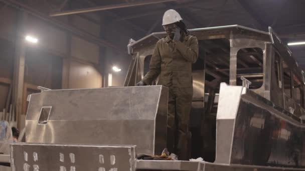 Full shot of female African dockyard worker wearing green overalls, hard hat and safety goggles, standing on stern of metal framework of boat, talking on smartphone - Footage, Video