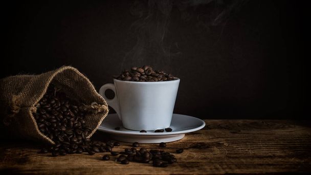 Άποψη των καβουρντισμένων σκουρόχρωμων κόκκων καφέ σε φλιτζάνι καφέ φρεσκοψημένα φασόλια σε τσάντα από γιούτα νεκρή φύση φωτογραφία - Φωτογραφία, εικόνα