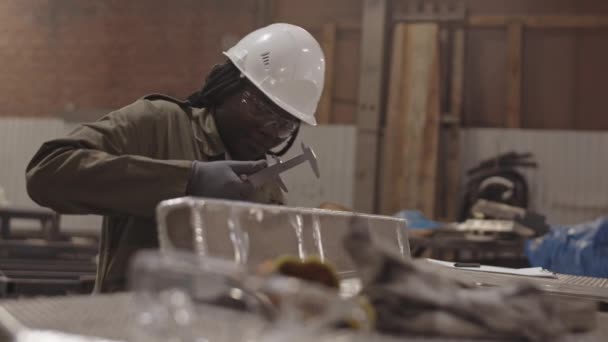 Gros plan moyen d'une travailleuse africaine portant un casque blanc et des lunettes de sécurité, utilisant des étriers mesurant la largeur de la pièce métallique, puis conciliant les dimensions sur le téléphone - Séquence, vidéo
