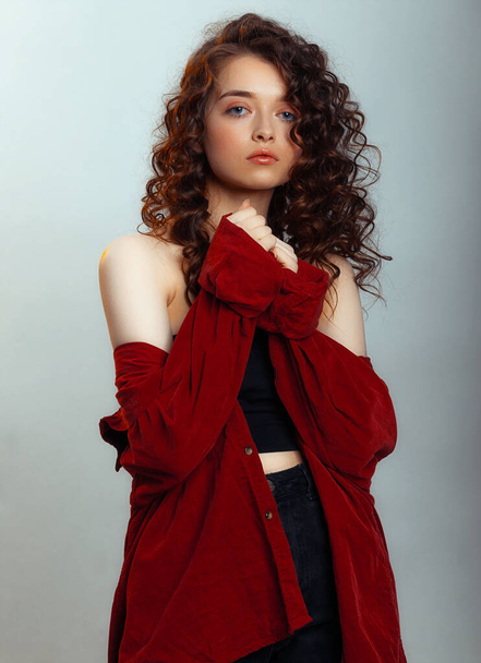 Attractive brunette with long curly hair. Female fashion model with wavy hairstyle in a red shirt posing on a gray background - Fotó, kép