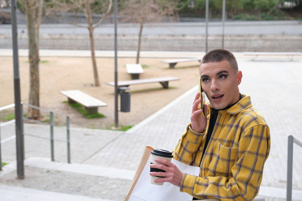 Jeune non binaire souriant et parlant sur le smartphone avec des sacs à provisions et une tasse de café. Maquiller une personne androgyne. - Photo, image