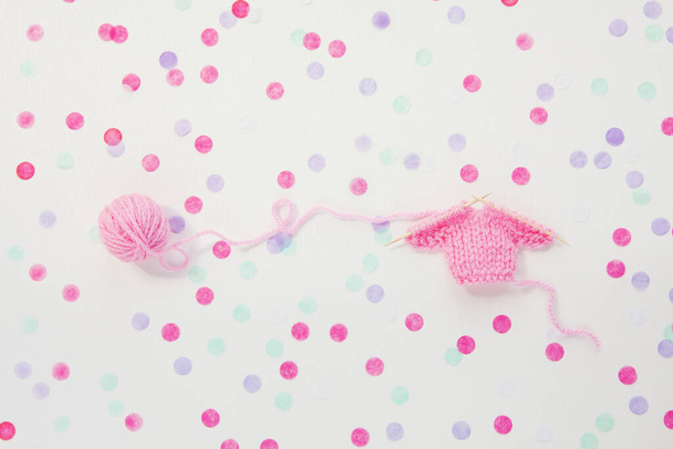 Knitted miniature pink sweater, round skein of thread, wooden needles on background of colorful round confetti. Concept gender party, boy or girl. Gift. Handmade. Copy space - Photo, Image