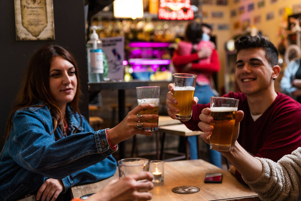Young people drink a toast with beer in a pub. - Detail of hands while toasting with glasses of beer - Happy people at a bar party after the harvest - Concept of friendship - Photo, Image
