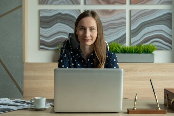Egy inges fiatal nő portréja, aki egy laptopon dolgozik az irodában, és a kamerába néz. - Fotó, kép