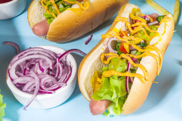 Fast food alla griglia, vari hot dog americani tradizionali con salsiccia, senape gialla, ketchup e insalata di verdure fresche, vista dall'alto su sfondo blu brillante con patatine fritte - Foto, immagini