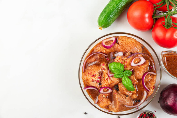 Marinated pork meat with fresh vegetables, bbq sauce, pepper, onion and basil in a bowl on white background. Top view. Marinating meat for cooking barbecue - Photo, image