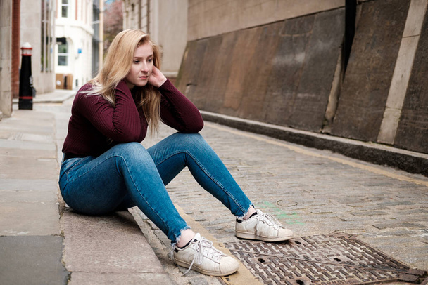 Portret van een jonge vrouw die op de vloer zit en er melancholisch uitziet. Ze draagt een kastanjebruine trui en een blauwe jeans. De straat is stenen paviljoen. - Foto, afbeelding