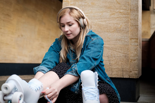 Young blonde woman sitting on the floor tying shoe laces on her roller skate. She is leaning against a wall and she is listening music on headphones. - 写真・画像