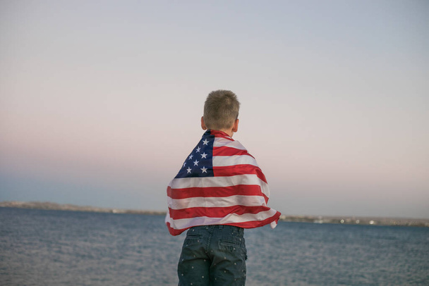 Маленький мальчик позволяет американскому флагу развеваться на ветру у моря. Патриотическая семья празднует День независимости США 4 июля. - Фото, изображение