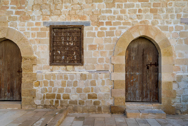 Две выветрившиеся деревянные арочные двери и закрытые ржавые кованые окна на кирпичной каменной стене - Фото, изображение