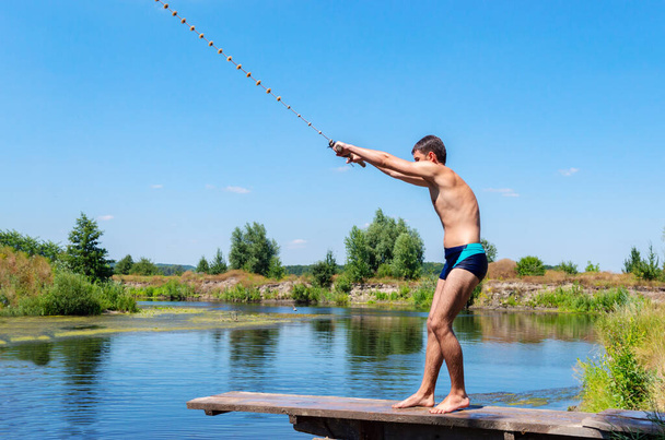 Un adolescent se tient sur un pont en bois et tient une corde oscillante, prêt à sauter dans la rivière par une journée d'été ensoleillée. Beau paysage, espace de copie. Concept de mode de vie sain - Photo, image