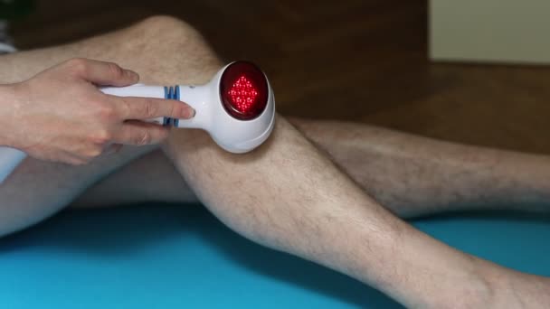 L'uomo massaggia il ginocchio malato con un massaggiatore elettrico con una luce rossa di calore - Filmati, video