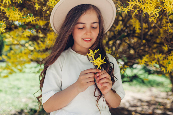 Stylowa mała uśmiechnięta dziewczynka trzymająca żółte kwiaty pod kwitnącym krzewem forsycji w wiosennym ogrodzie. Dzieciak w kapeluszu - Zdjęcie, obraz