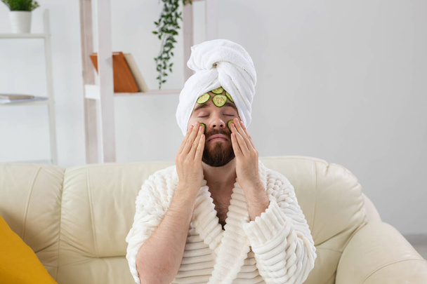 Baard man geniet met een cosmetisch masker op zijn gezicht gemaakt van plakjes komkommer. Mannen huidverzorging en spa thuis concept - Foto, afbeelding