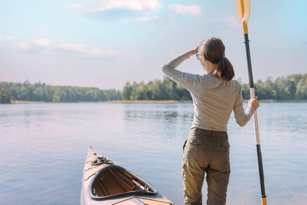 A young woman next to a kayak stands with a paddle and looks into the distance. - Fotografie, Obrázek