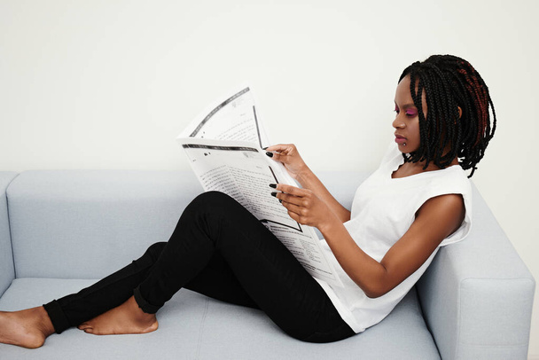 Αφροαμερικανό κορίτσι που διαβάζει εφημερίδα στον καναπέ - Φωτογραφία, εικόνα