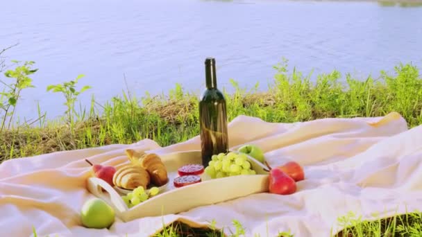 Nehir kenarında piknik. Açık renkli bir yatak örtüsü, içinde meyve, kek ve bir şişe şarap olan ahşap bir tepsi. Batan güneşin ışığı, hoş sakin bir atmosfer, parlak renkler.. - Video, Çekim