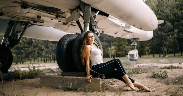 beautiful young brunette girl stands on the background of old military aircraft. Girl in a white shirt and black pants in nature. Military equipment. Portrait half-length. girl posing - Foto, immagini