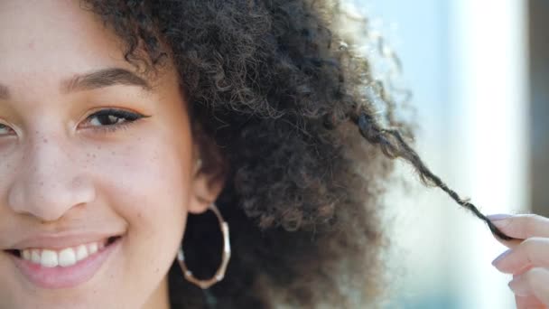 Hermosa joven africana confiada se ríe. Estudiante étnico americano feliz, peinado afro rizado, pendientes perforados redondos, maquillaje ligero y sonrisas pecas y muestra sus dientes blancos saludables - Imágenes, Vídeo