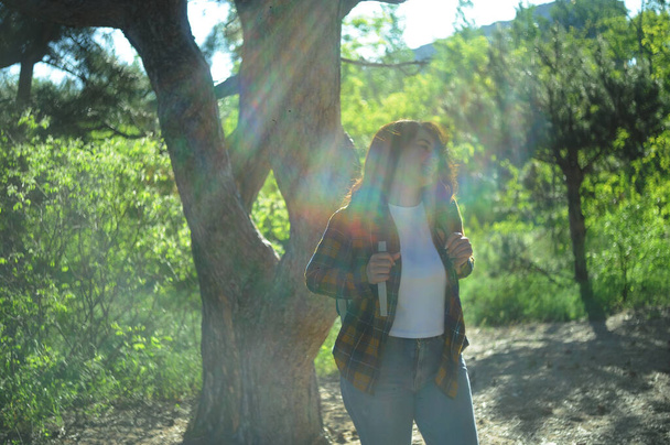 Hymyilevä onnellinen nainen keltaisessa ruudullisessa paidassa matkakassin kanssa viettää aikaa metsässä. Kesälomien käsite.  - Valokuva, kuva