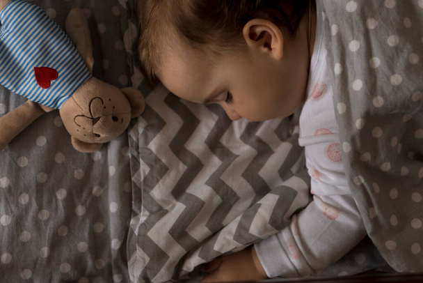 トップ表示かわいいぽっちゃりした赤ちゃんの女の子の子供は、自宅でテディベアと白とピンクのパジャマで昼休みにグレーのベビーベッドで甘く寝ています。小児期,レジャー,快適さ,医学,健康の概念 - 写真・画像