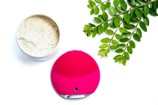 розовая электронная щетка для чистки лица, скраб для лица и зеленые листья на белом фоне - Фото, изображение