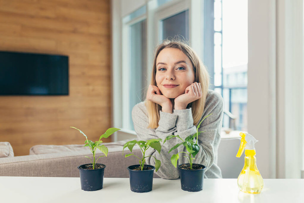 Young beautiful blonde woman, at home takes care of flower pots, pours flowers sitting at the table - 写真・画像
