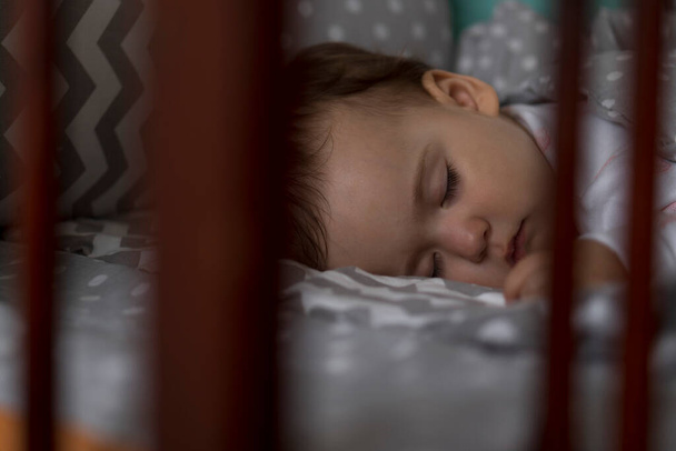 Bonito alegre pequena menina gordinha dormindo docemente no berço do bebê cinza durante o tempo de almoço em pijama branco e rosa com ursinho de pelúcia em casa. Infância, lazer, conforto, medicina, conceito de saúde - Foto, Imagem