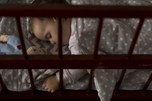 Cute cheerful little chubby baby girl sleeping sweetly in grey baby crib during lunch rest time in white and pink pajamas with teddy bear at home. Childhood, leisure, comfort, medicine, health concept - Photo, Image
