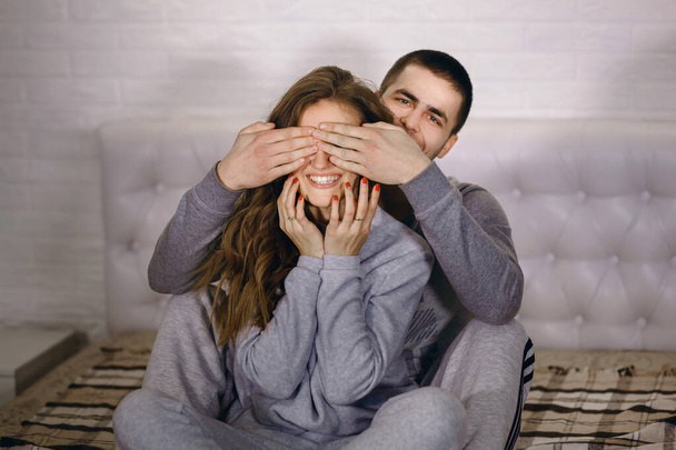 Młoda para siedzi na łóżku i dobrze się bawi. Młody mężczyzna zakrywa oczy swojej kobiety dłońmi. Facet i dziewczyna są ubrani w domowe ciuchy. Szczęśliwy związek miłości - Zdjęcie, obraz
