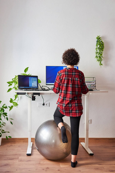 Onherkenbare vrouw telewerken aan een verstelbaar bureau met een knie rustend op een fitbal - Foto, afbeelding