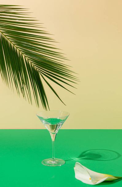 Вид сверху на листья пальмы и бокал мартини на зеленом и песочном фоне. Летний напиток минимальная концепция. Элегантная композиция. - Фото, изображение