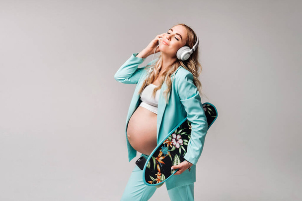 A pregnant girl in a turquoise suit with a skateboard in her hands and headphones stands on a gray background. - Foto, Imagen
