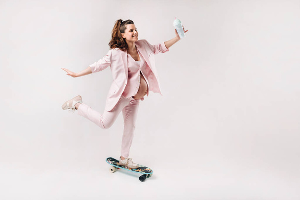 A pregnant girl in a pink suit with a skateboard in her hands stands on a gray background. - Fotografie, Obrázek