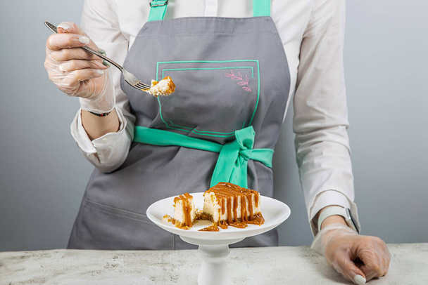 A pastry chef breaks off a piece of the freshest cheesecake drizzled with caramel with a fork on a white plate - Φωτογραφία, εικόνα
