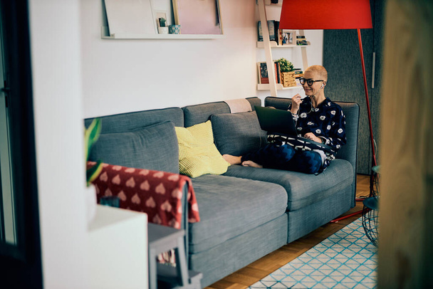 Viehättävä vanhempi liikenainen töissä kotoa. Eläkeläinen käyttää kannettavaa tietokonetta istuessaan sohvalla kotona. - Valokuva, kuva