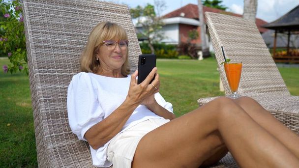 Woman over 50 on a sunbed smiling at her smartphone while video calling with beloved ones. Staying connected on a long distance.  - Fotografie, Obrázek